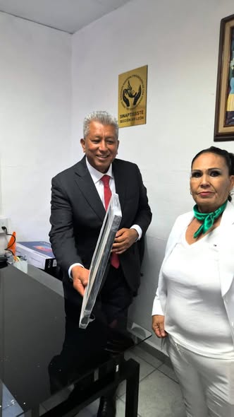 Dos integrantes posan en la oficina de la Sección XVI junto a un reconocimiento enmarcado.
